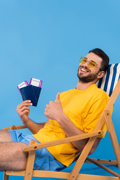 Веселий чоловік в сонцезахисних окулярах показує, як і тримає паспорти з авіаквитками на стільці на синьому фоні
  - Фото, зображення