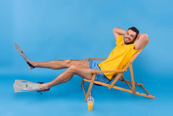 Pozitivní muž v plavání ploutve sedí na palubě židle v blízkosti pomerančové šťávy na modrém pozadí  - Fotografie, Obrázek