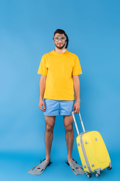 Молодой человек в плавательных очках и ластах держит чемодан на синем фоне  - Фото, изображение