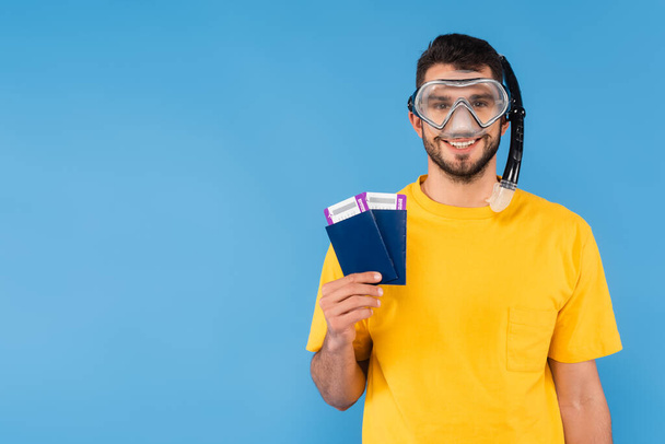 Hombre positivo en gafas de natación sosteniendo boletos aéreos y pasaportes aislados en azul  - Foto, Imagen