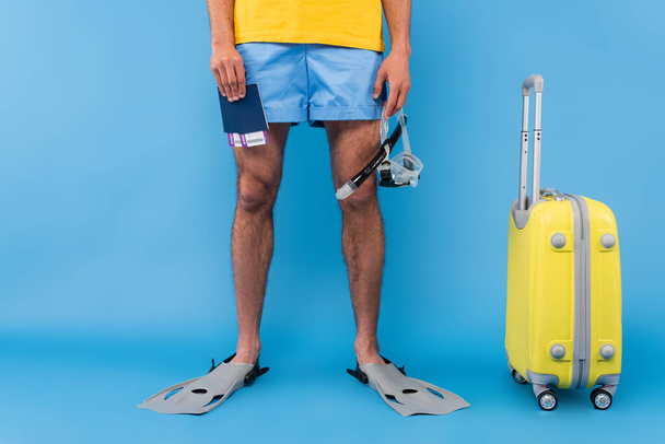 Vista recortada del hombre en aletas de natación con pasaportes y gafas cerca de la maleta sobre fondo azul  - Foto, Imagen