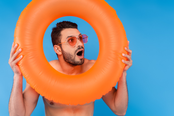 Uomo eccitato in occhiali da sole tenendo anello gonfiabile isolato su blu  - Foto, immagini