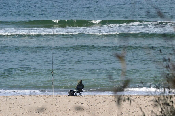 il pescatore è seduto sulla spiaggia accanto alla canna da pesca, chiedendosi se prenderà qualcosa  - Foto, immagini