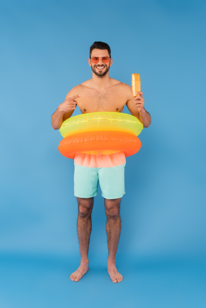 Hombre sin camisa en anillos inflables apuntando al protector solar sobre fondo azul  - Foto, Imagen