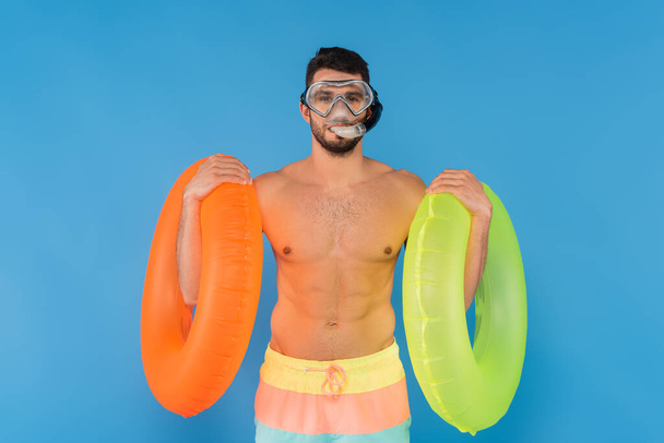 青い上に隔離されたインフレータブルリングを保持している水泳ゴーグルの恥知らずな男 - 写真・画像