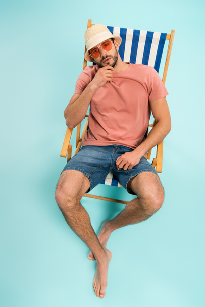 High angle view of bearded man in straw hat and sunglasses sitting on deck chair on blue background  - Fotoğraf, Görsel