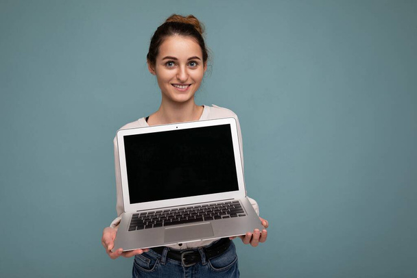 Όμορφη χαμογελαστή νεαρή γυναίκα κρατώντας υπολογιστή netbook κοιτάζοντας κάμερα φορώντας λευκό πουκάμισο απομονωμένο σε μπλε φόντο - Φωτογραφία, εικόνα