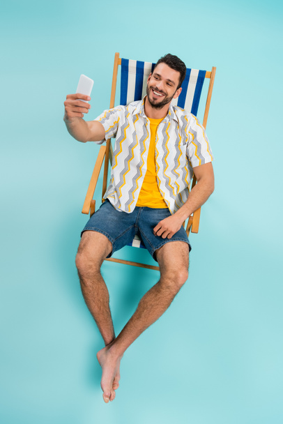 Hoge hoek uitzicht van lachende man nemen selfie terwijl zitten op dek stoel op blauwe achtergrond  - Foto, afbeelding