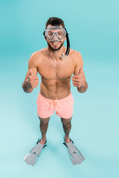 Hoge hoek uitzicht van shirtloze man in zwembril en flippers tonen als op blauwe achtergrond  - Foto, afbeelding