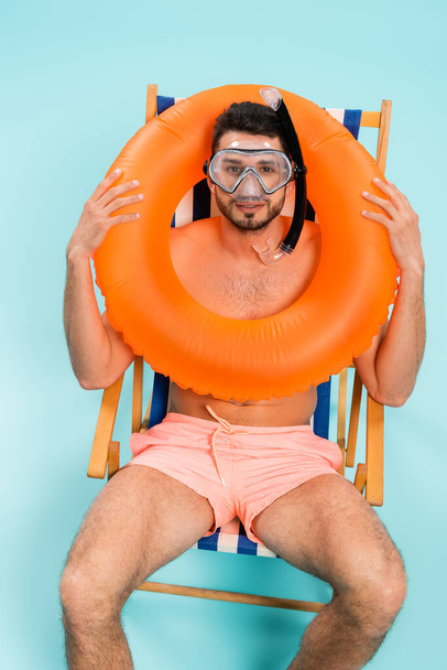 High angle view of smiling man in swimming goggles holding inflatable ring on blue background  - Φωτογραφία, εικόνα
