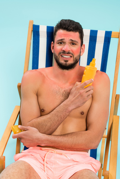 Besorgter Mann mit Sonnenbrand hält Sonnencreme auf Liegestuhl isoliert auf blauem Grund  - Foto, Bild