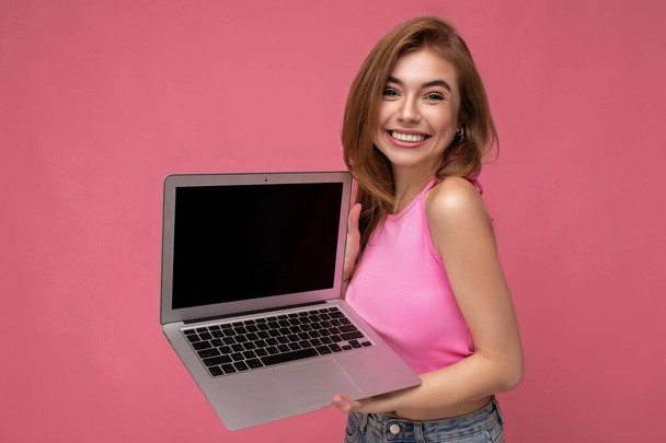 Foto de una hermosa joven sosteniendo computadora portátil mirando a la cámara aislada sobre un fondo colorido - Foto, imagen
