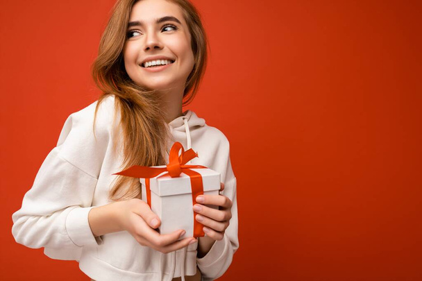 Shot of attractive positive smiling young blonde woman isolated over colourful background wall wearing everyday trendy outfit holding gift box and looking to the side - Photo, Image