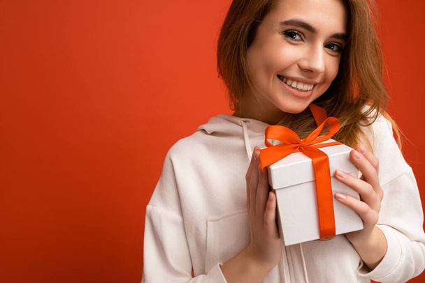Closeup photo shot of acharming sexy positive smiling blonde young woman isolated over colorful background wall wearing white hoodie holding white gift box with red ribbon and looking at camera - Fotó, kép