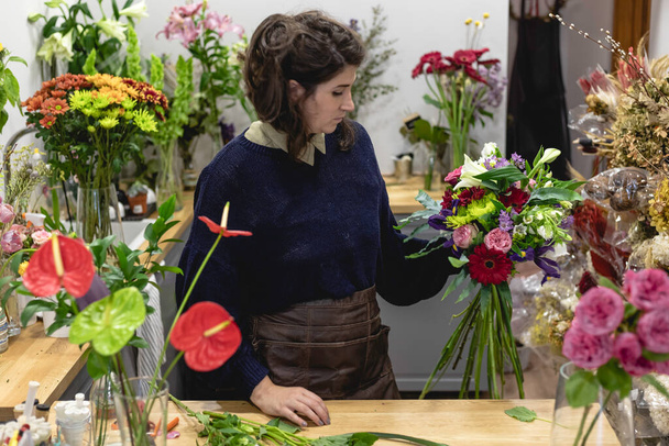 Άποψη μιας νεαρής γυναίκας που εργάζεται ως ανθοπώλης και δημιουργεί ένα μπουκέτο με πολύχρωμα λουλούδια στο κατάστημά της, γυναίκα επιχειρηματίας έννοια. - Φωτογραφία, εικόνα