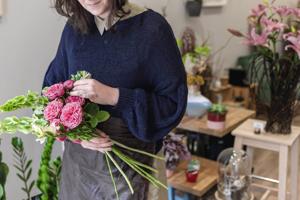 Άποψη μιας γυναίκας που σχεδιάζει ένα μπουκέτο λουλουδιών με πράσινα και μοβ λουλούδια στο ανθοπωλείο της φορώντας μια ποδιά, οι νέες γυναίκες επιχειρηματίες έννοια. - Φωτογραφία, εικόνα