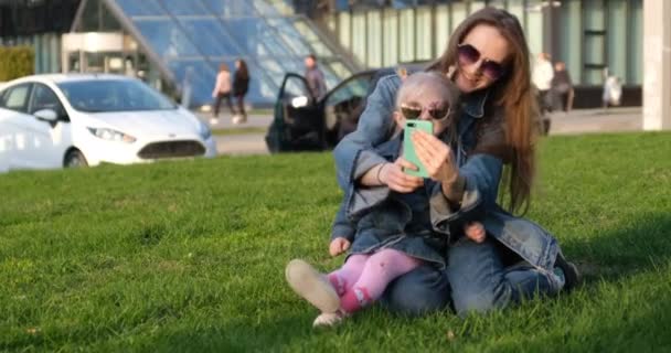 Mam en dochtertje praten via een videolink op het gazon. Een vrouw en een meisje nemen een selfie op hun telefoon. Familie ontspant op het gazon in de stad - Video