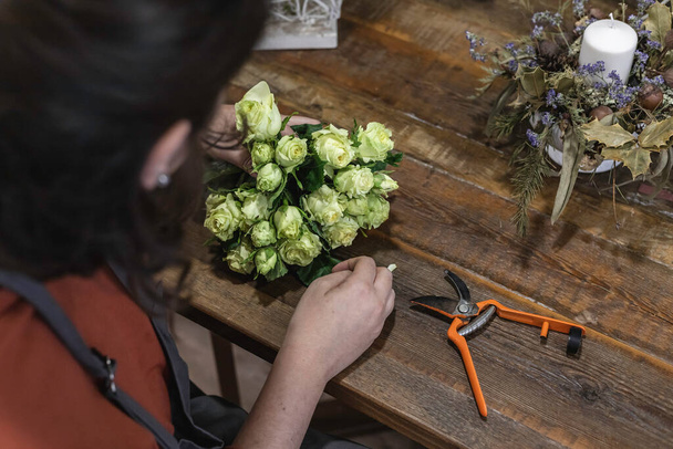 Вид молодой женщины, работающей флористом в цветочном магазине, сидящей за деревянным столом и создающей цветочные украшения, концепция бизнесвумен. - Фото, изображение