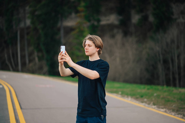 Knappe man in donkere kleren loopt over een bergweg in het bos en maakt foto 's op een smartphone camera. Toerist maakt foto 's van de soort tijdens het wandelen. - Foto, afbeelding