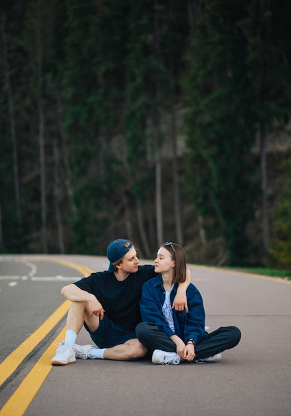 Hombre y mujer en ropa casual descansando sentados en la carretera en las montañas y abrazándose sobre un fondo de bosque de coníferas. Vertical. Ocio en la naturaleza. - Foto, imagen