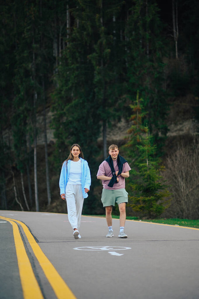 Stylish young couple man and woman in a fork in the mountains, walking on the road on a background of coniferous trees. Paparazzi photo of a beautiful couple on vacation in the woods. - Foto, Imagen