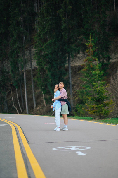 Lächelndes Paar in stylischer Freizeitkleidung, das auf einer Bergstraße in den Armen steht und mit einem Lächeln im Gesicht für die Kamera posiert. Vertikal - Foto, Bild