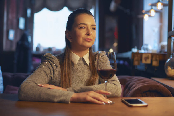A beautiful young girl sits at a wooden table in the premises of a wine bar and drinks wine from a glass. - Фото, изображение