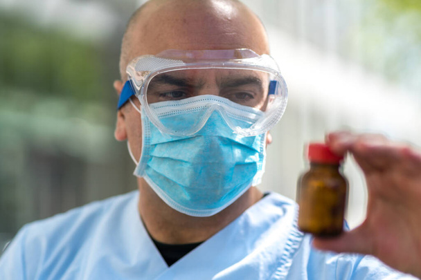 Από εστιασμένο γιατρό φορώντας εργαστηριακή ποδιά, χειρουργική μάσκα και γυαλιά ιατρικής ασφάλειας προσφέροντας ένα μπουκάλι ή βάζο με υγρά αντιβιοτικά. Επιλεκτική εστίαση - Φωτογραφία, εικόνα