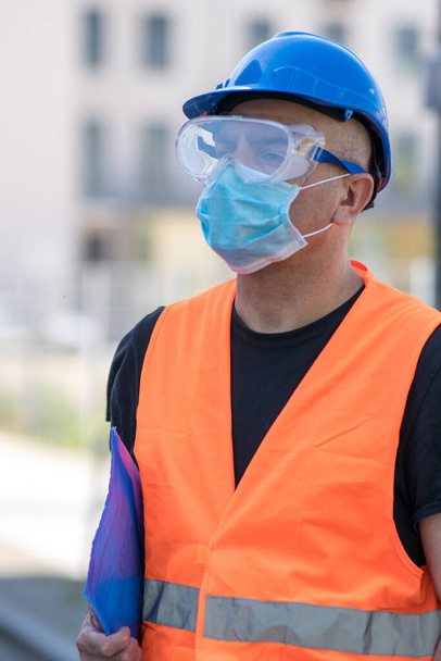 Безопасность и охрана труда от неблагоприятных условий труда. Строитель в синей каске, светоотражающий жилет и защитная хирургическая маска - Фото, изображение