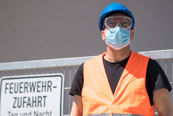 Seguridad y salud en el trabajo y protección contra las condiciones adversas en el trabajo. Trabajador de la construcción con sombrero duro azul, chaleco reflectante y máscara quirúrgica protectora - Foto, Imagen