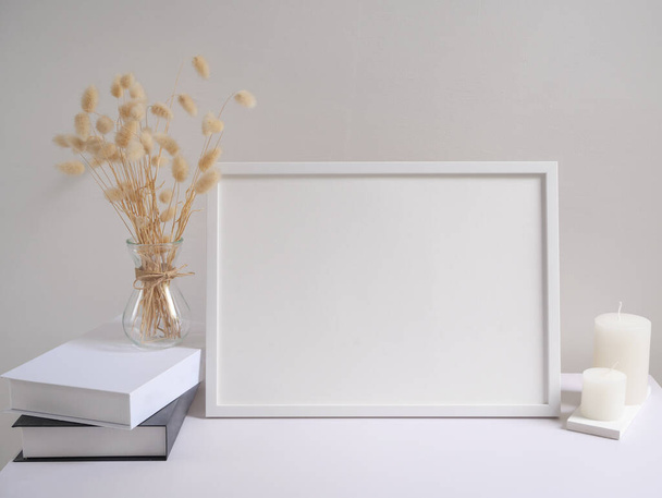 Mock up poster frame zwart-wit boeken gedroogd Lagurus ovatus bloemen samenstelling in modern glas vaas en kaarsen op witte tafel en cement muur achtergrond  - Foto, afbeelding