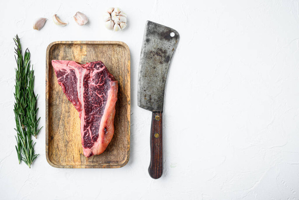 Çiğ mermer biftek seti, T kemik kesimi, eski kasap bıçağı, beyaz taş arka planda, üst görünüm düz, metin için kopyalama alanı - Fotoğraf, Görsel