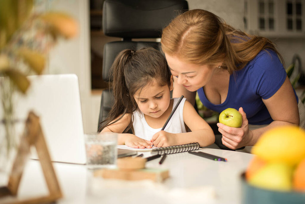 Mother and daughter sitting at desk in living room and studying online together - 写真・画像