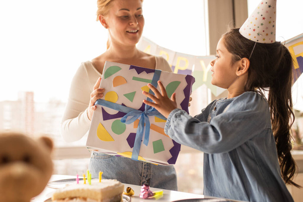 Woman giving a gift to her daughter wearing party cap while spending her birthday - Foto, afbeelding