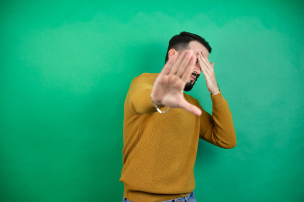 Pohledný muž v neformálním oblečení na izolovaném zeleném pozadí, zakrývajícím oči rukama a zastavujícím gestem se smutným a ustrašeným výrazem. Ztrapněný a negativní koncept. - Fotografie, Obrázek