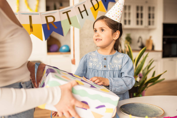 Girl wearing party hat looking at her mother while accepting her birthday gift - Foto, immagini