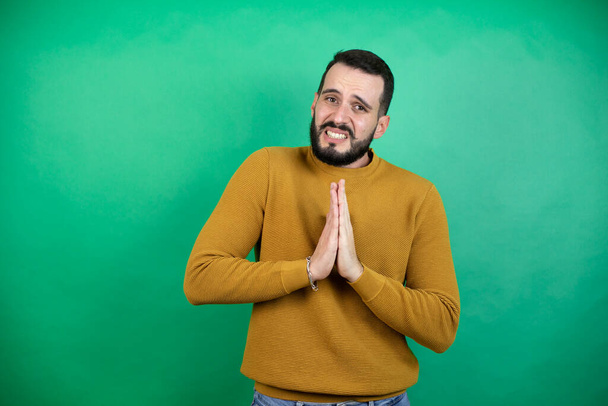 Красивий чоловік в повсякденному одязі над ізольованим зеленим тлом молиться і молиться руками разом з виразом надії на обличчі дуже емоційно і турбується
 - Фото, зображення