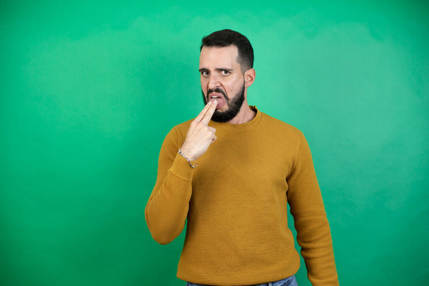 Όμορφος άντρας που φοράει καθημερινά ρούχα πάνω από απομονωμένο πράσινο φόντο αηδιασμένος με το χέρι του μέσα στο στόμα του. - Φωτογραφία, εικόνα