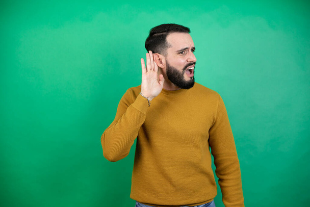 Jóképű férfi alkalmi ruhát visel elszigetelt zöld háttér meglepett kézről fülre hallgat egy meghallgatás pletykák vagy pletykák - Fotó, kép