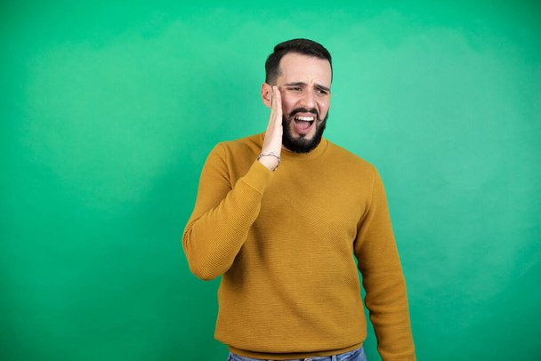 Knappe man draagt casual kleding over geïsoleerde groene achtergrond aanraken mond met de hand met pijnlijke uitdrukking als gevolg van kiespijn of tandheelkundige ziekte op tanden - Foto, afbeelding