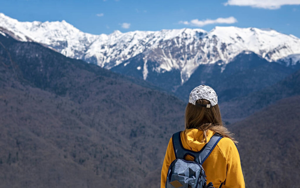 Zadní pohled Mladá blondýnka ve žluté mikině s kapucí a batohem na pozadí zasněžených Kavkazských hor. Turistika - Fotografie, Obrázek