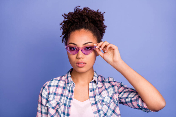Foto de dama joven y confiada seria vestida con camisa a cuadros brazo gafas de color rosa aislado color púrpura fondo - Foto, Imagen