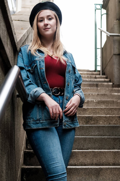 Młoda blondynka pozująca na schodach w berecie, dżinsowej kurtce i spodniach oraz czerwonej koszulce. - Zdjęcie, obraz