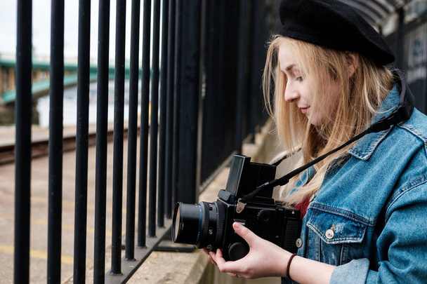 Jonge vrouw maakt foto 's met een middelgrote filmcamera. Ze draagt een spijkerjack en een zwarte baret. Naar de camera kijken. - Foto, afbeelding