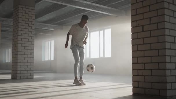 Een zwarte man traint met een voetbal op een ondergrondse parkeerplaats in het zonlicht in slow motion. Jongleren met de bal, de pas trainen en schoppen. Werken aan succes - Video