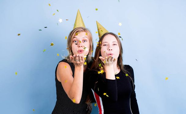 Doğum günü partisi. Doğum günü şapkalı iki genç kız ellerinde balonlarla mavi arka planda doğum günü kutluyorlar, konfeti üflüyorlar. - Fotoğraf, Görsel
