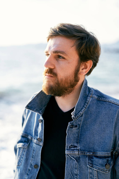 Portret zamyślonego mężczyzny w jeansowej kurtce spoglądającego w dal na tle morza. Zbliżenie - Zdjęcie, obraz