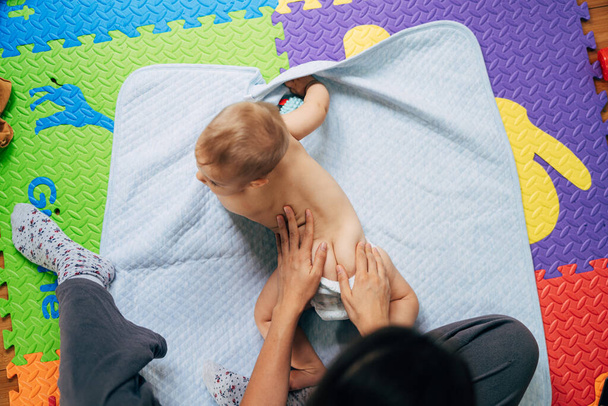 Το μωρό με τις πάνες ξαπλώνει στην κοιλιά του σε μια μπλε κουβέρτα πάνω από ένα χρωματιστό χαλί στο πάτωμα. Η μαμά έβαλε τα χέρια της στο μωρό στην πλάτη. - Φωτογραφία, εικόνα