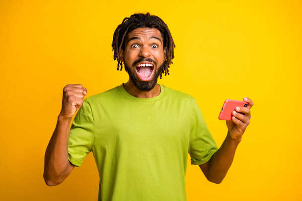 Photo of young man happy excited wow celebrate win victory goal hold smartphone wear green t-shirt isolated over yellow color background - Photo, Image