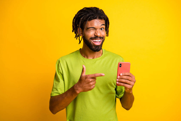 Photo of excited dark skin guy recommend social media ads point smartphone wear t-shirt isolated over shine color background - Foto, imagen